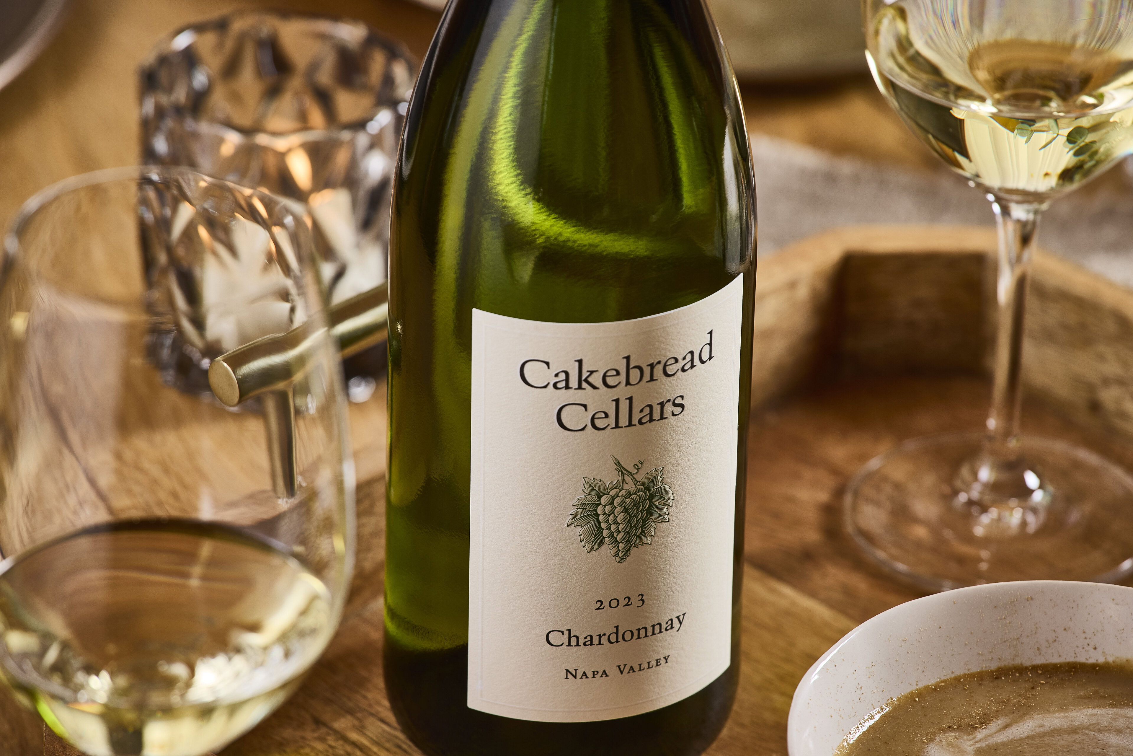 A lush green bottle of Cakebread Cellars Chardonnay wine on a wooden table with wine glasses.
