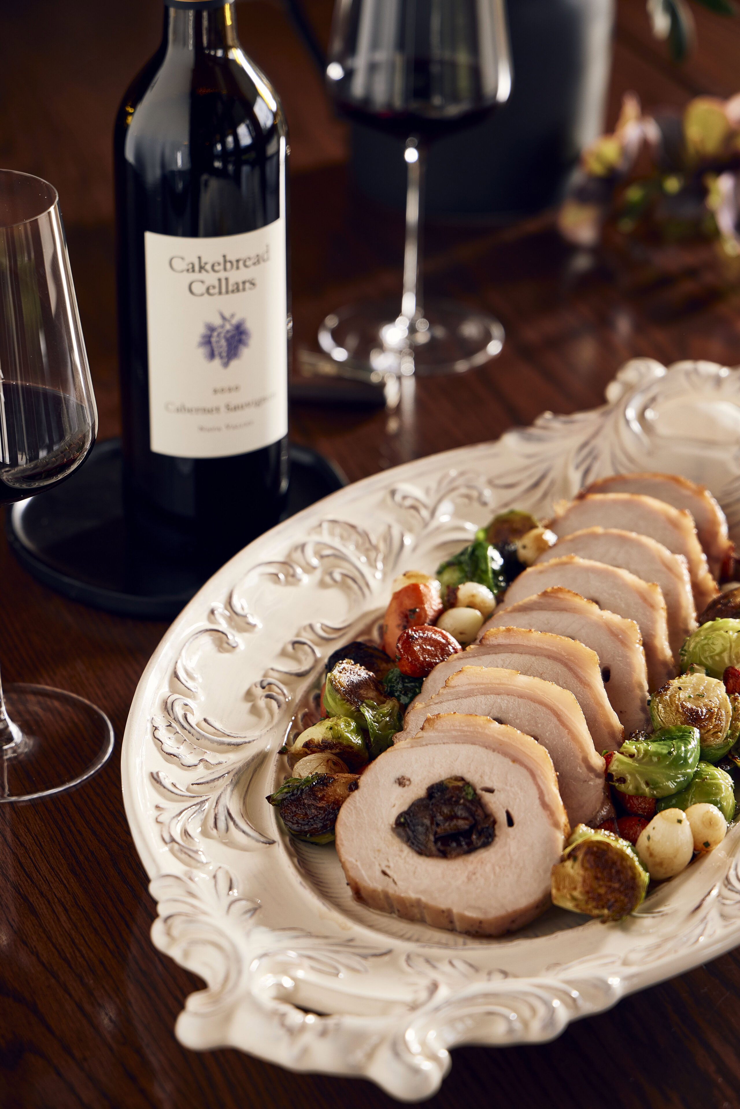 A lush pork loin stuffed with French prunes and pecans, sliced and served on an ornate white platter with roasted Brussels sp.