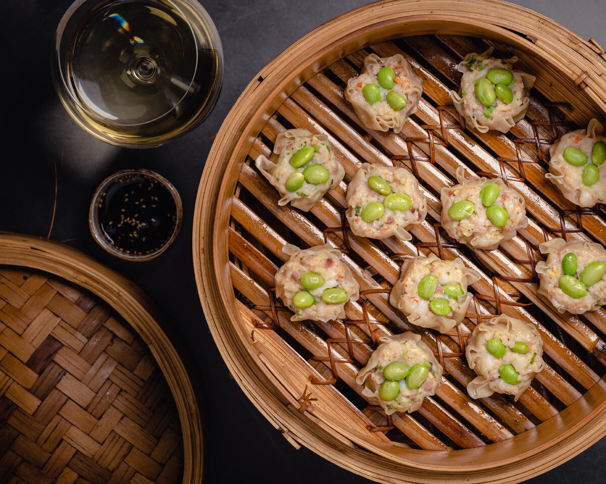 Delicate steamed dumplings topped with green edamame beans served in a traditional bamboo steamer basket.