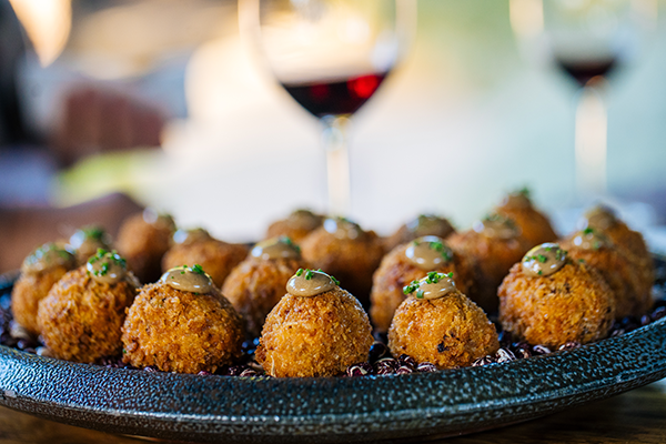 Crispy wild mushroom arancini served on a black platter with green herb garnish, paired with glasses of red wine.