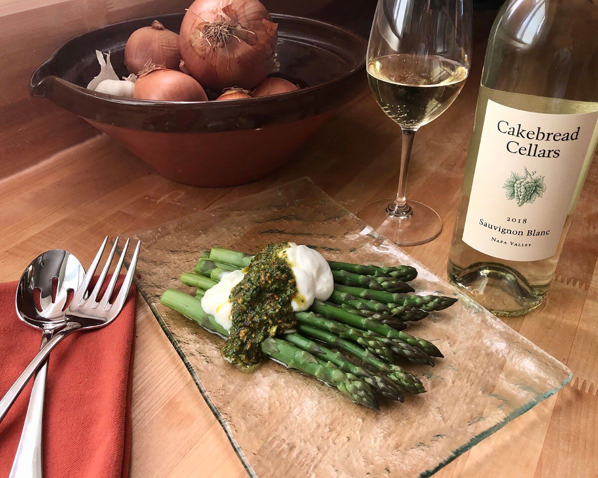 A lush plate of fresh asparagus topped with burrata cheese and pistachio salsa verde, served alongside a glass of white wine.