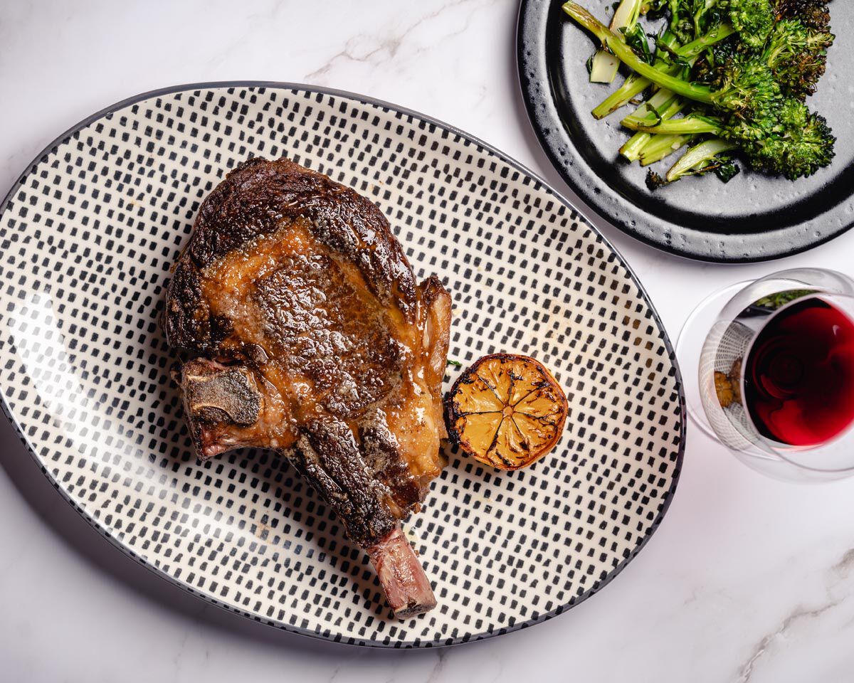 Seared bone-in ribeye steak with roasted broccolini and a glass of red wine on a white marble surface.
