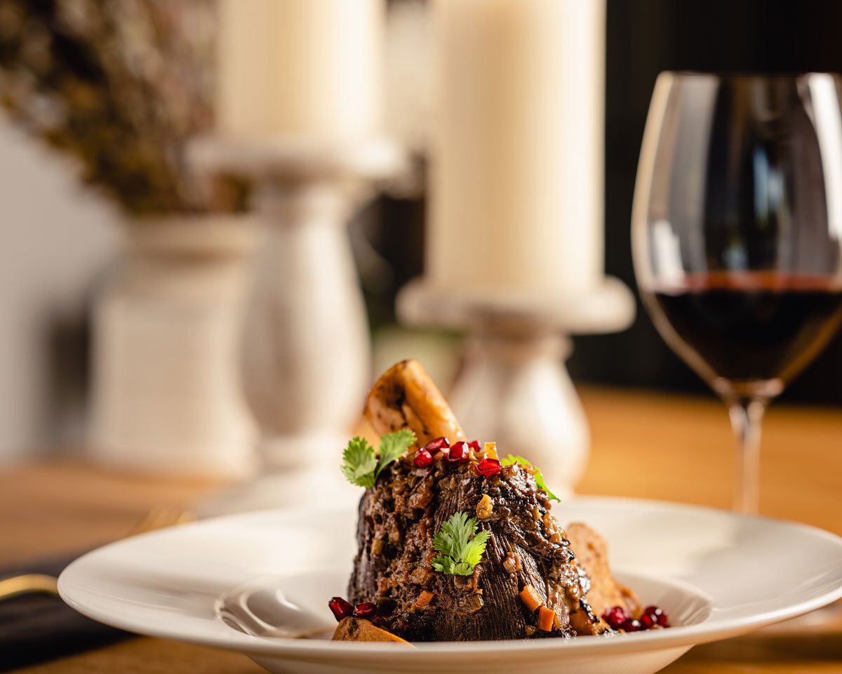 Braised venison shank garnished with herbs and pomegranate seeds, served with a glass of red wine on a white plate.
