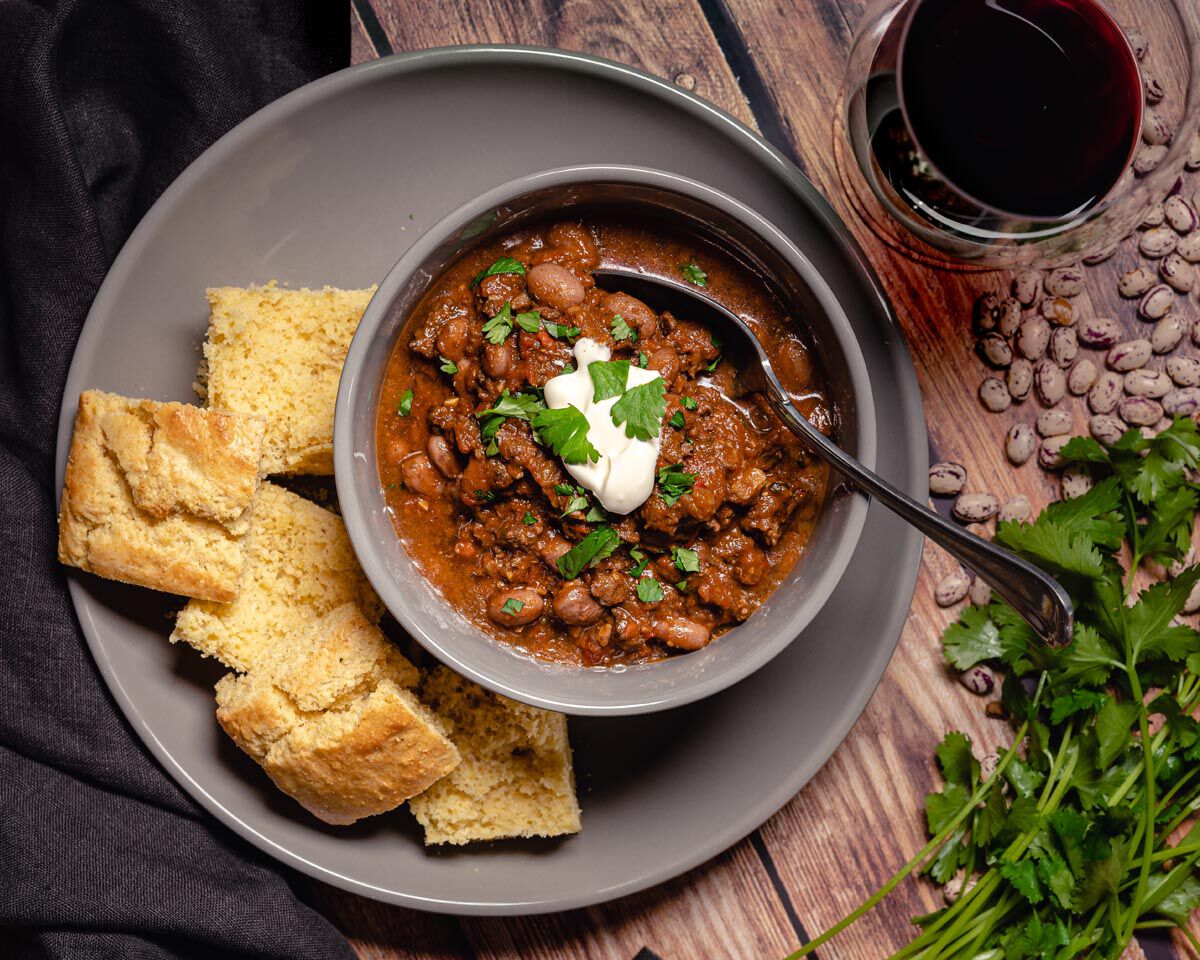 Hearty venison chili served with cornbread and a glass of red wine on a rustic wooden table. Fresh cilantro garnishes the chi.