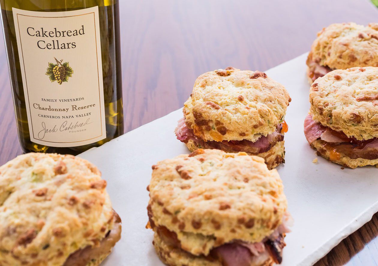Creamy Carmody cheese and green onion biscuits with smoked ham and red pepper jelly on a white serving board, alongside a bot.