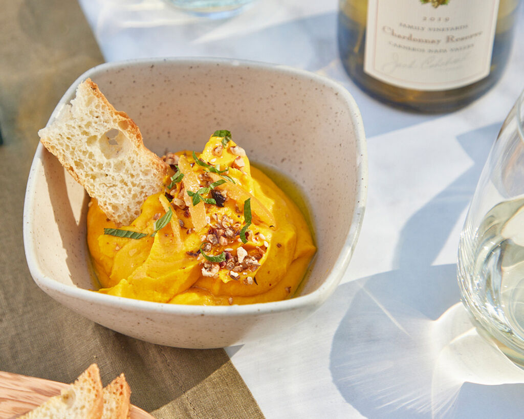 Creamy carrot hummus topped with chopped pecans and herbs, served with a slice of crusty bread in a white bowl.