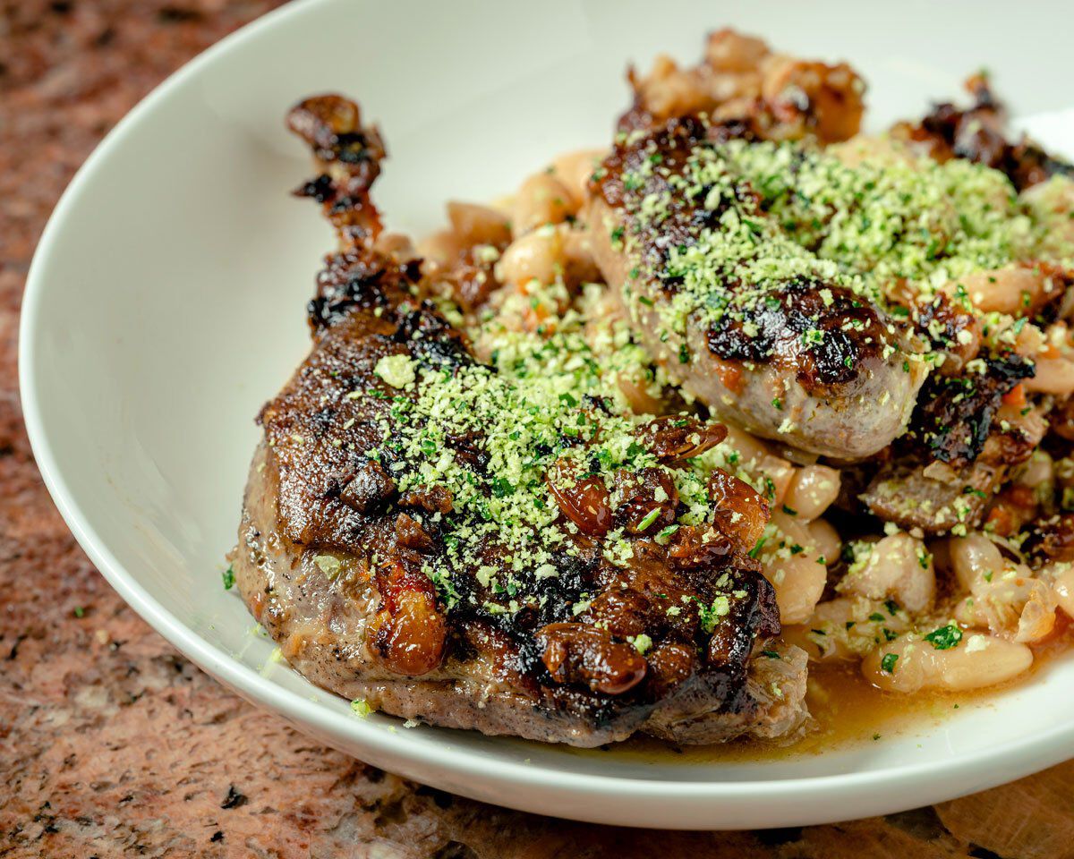 Roasted chicken thighs with beans and green herbs, served in a white bowl, highlighting a hearty cassoulet dish.