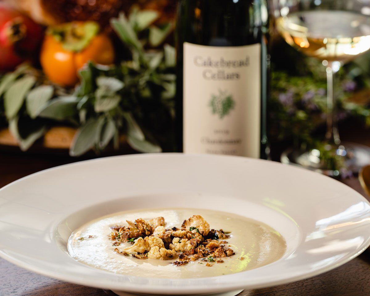 Creamy cauliflower soup garnished with pancetta and capers, served in a white bowl with a bottle of Cakebread Cellars wine in.