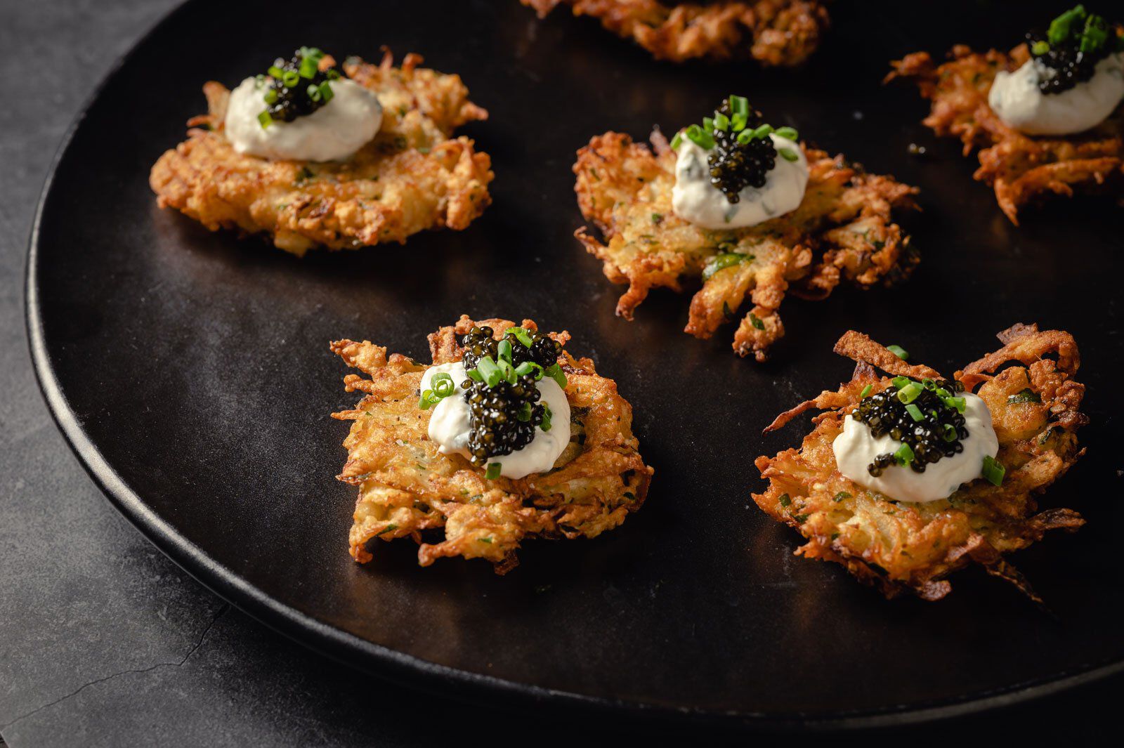 Crispy celery root latkes topped with horseradish sauce and caviar on a black plate, served as a gourmet appetizer.