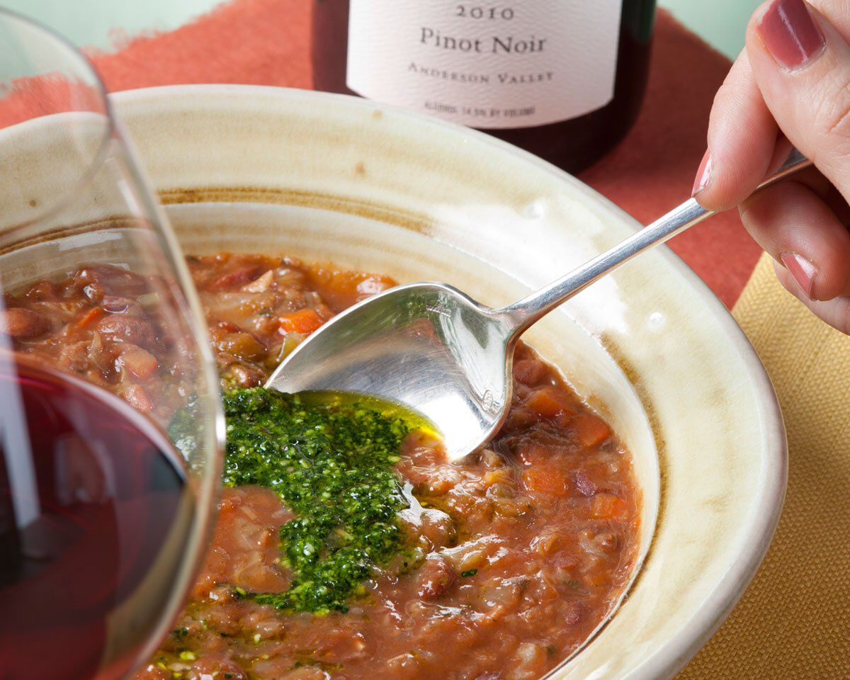Rich cranberry bean, cabbage, and red wine soup served in a rustic bowl with a spoon, featuring Cavolo Nero pasta and a glass.