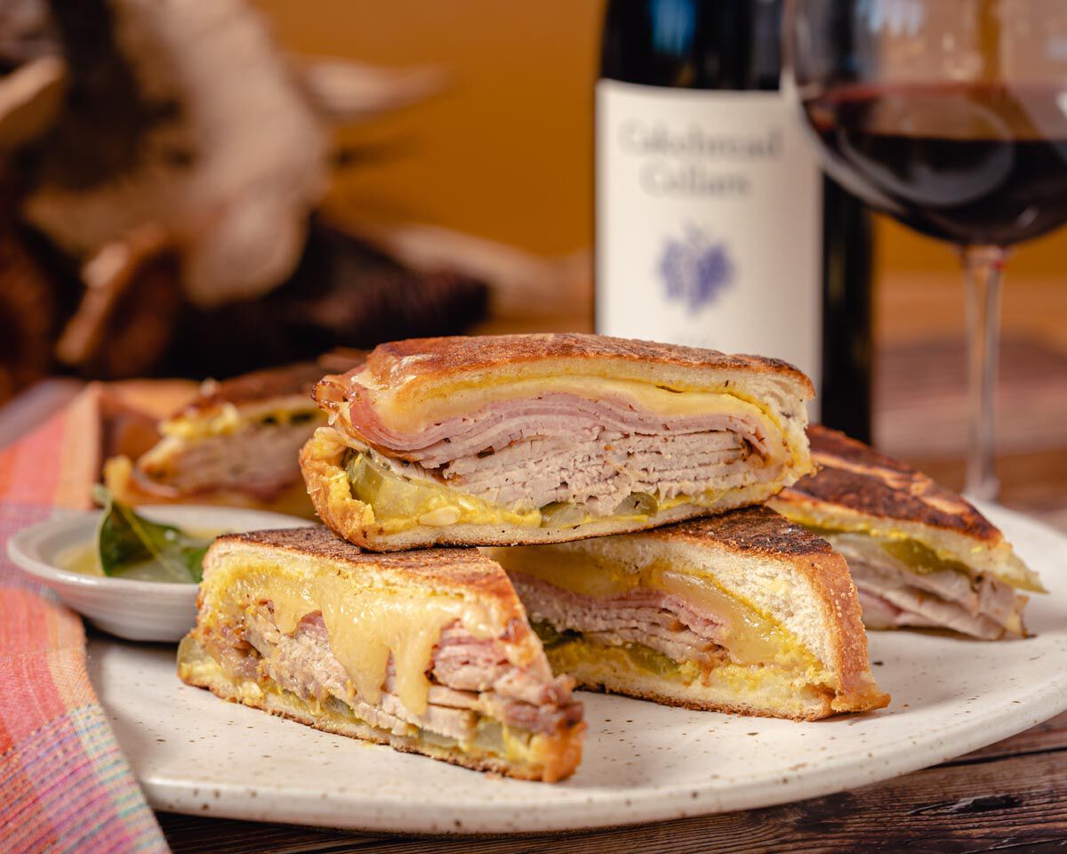 Grilled Cubano sandwich with ham, cheese, and pickles on a white plate, with a glass of red wine and a bottle of wine in the.