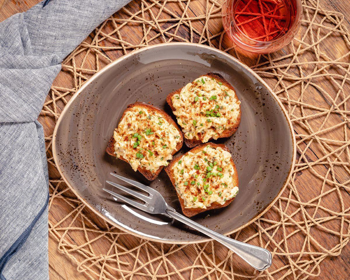 Creamy deviled crab toasts served on a rustic gray plate with a fork, garnished with herbs, and accompanied by a glass of ros.