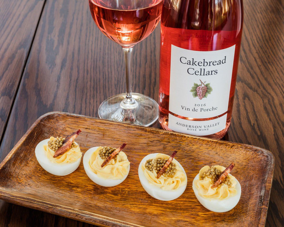 A colorful glass of rosé wine and a bottle of Cakebread Cellars wine on a wooden table with deviled eggs topped with pickled.