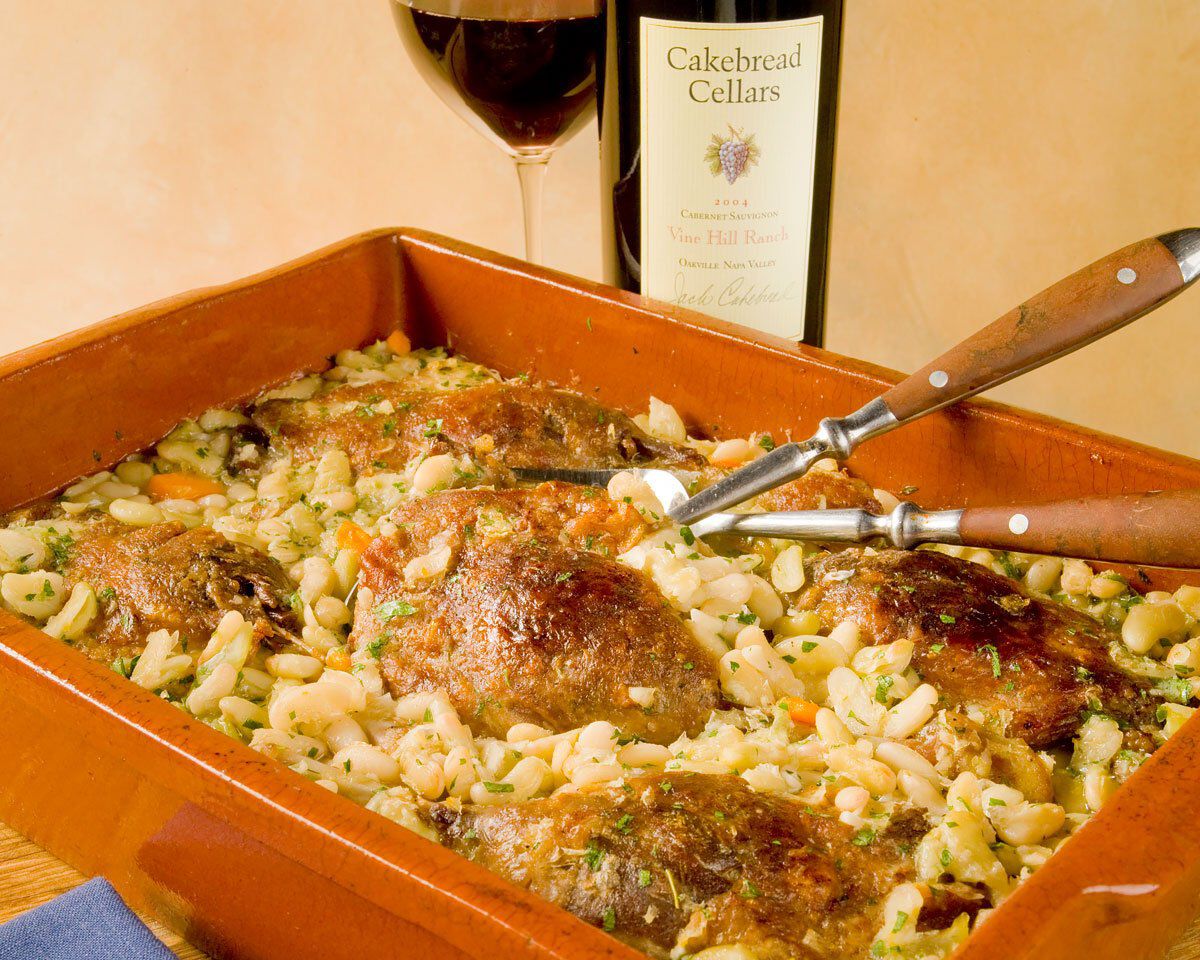 Roasted duck confit with pasta and vegetables in a ceramic baking dish, accompanied by a glass of red wine and a bottle of Ca.