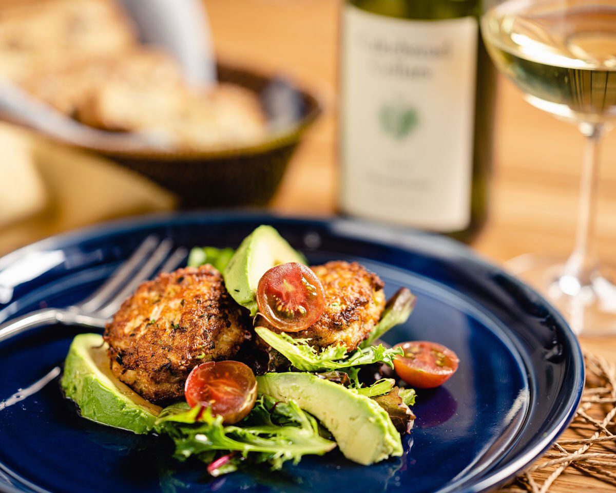 A colorful Dungeness crab cake served with fresh avocado slices and cherry tomatoes on a dark blue plate.