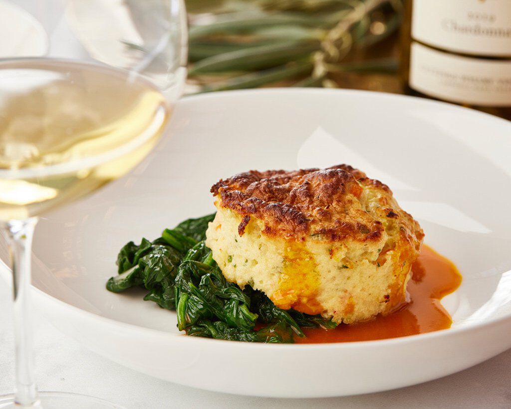 Creamy Dungeness Crab Soufflé served with sautéed greens and a glass of white wine on a white plate.