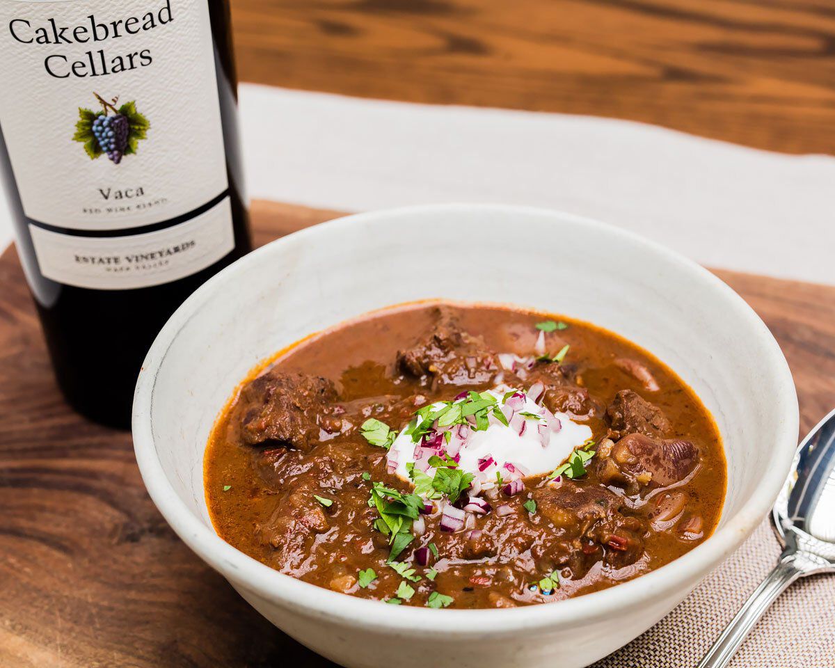 Elk chili with Mexican chocolate and lime crema in a white bowl, garnished with cilantro and red onion, served with a bottle.
