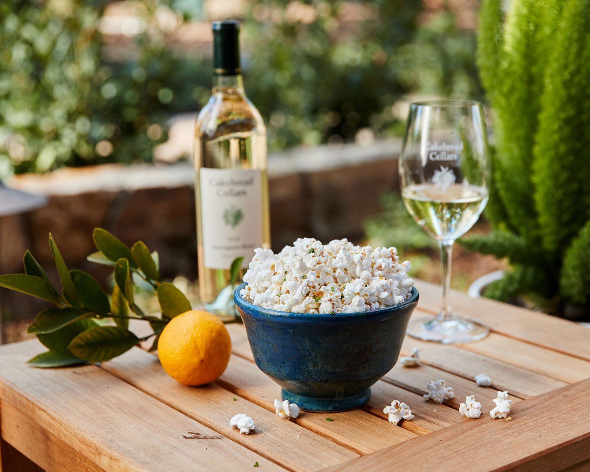 A lush outdoor setting with a bowl of popcorn flavored with Meyer lemon and Parmesan, a bottle of white wine, and a glass of.