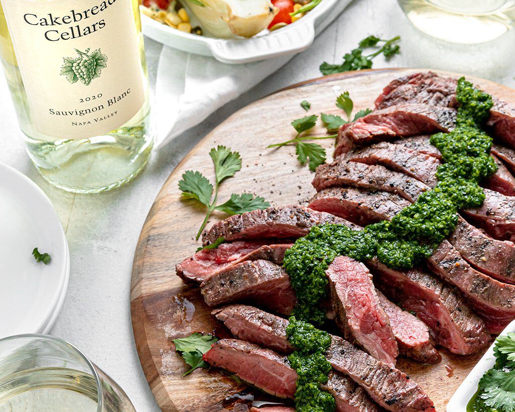 Grilled flat iron steak with chimichurri sauce on a wooden cutting board, garnished with fresh herbs, served with wine and sa.