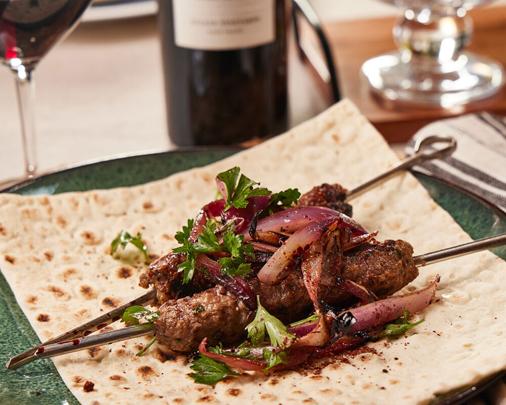 Grilled harissa-spiced lamb kebabs with red onion and herbs on flatbread, served with wine in a restaurant setting.