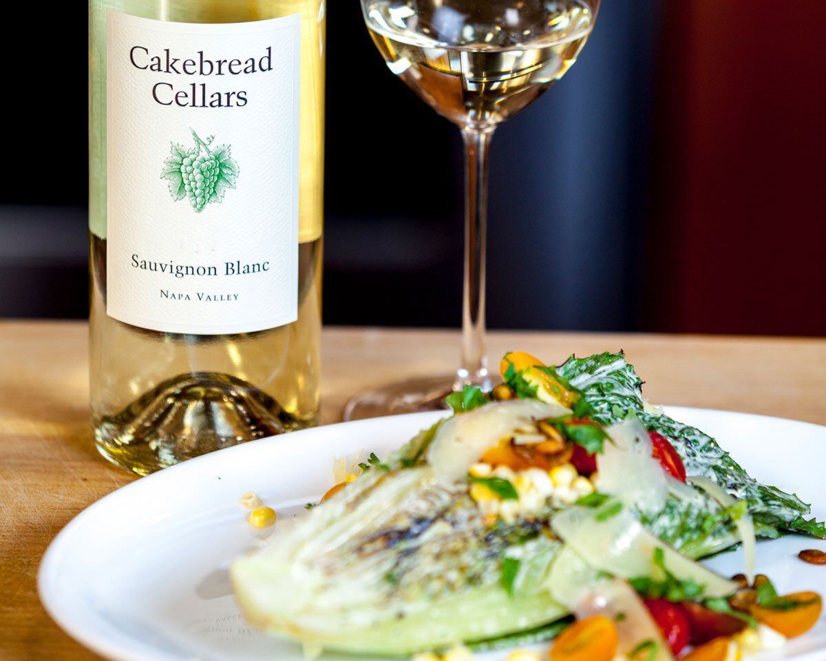 Bottle of Cakebread Cellars Sauvignon Blanc with a glass of white wine and a grilled romaine salad on a wooden table.
