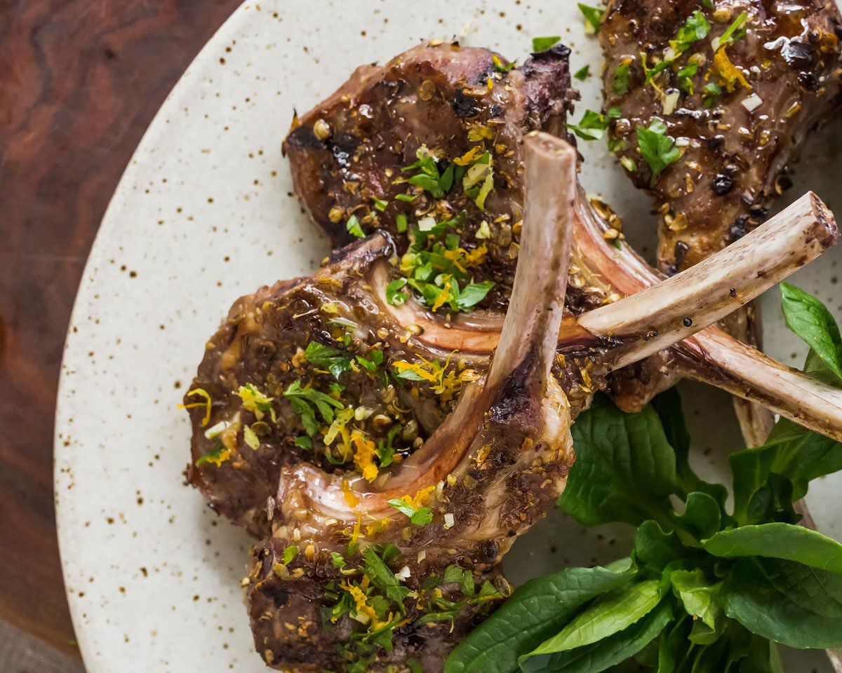 Succulent grilled herb-rubbed lamb chops garnished with fresh herbs on a white plate. Perfect pairing with Cakebread Cellars.