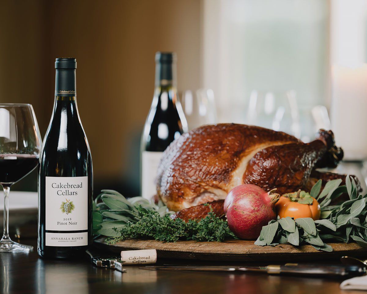 Bottles of Cakebread Cellars Pinot Noir with a roasted smoked turkey and fresh vegetables on a dining table.