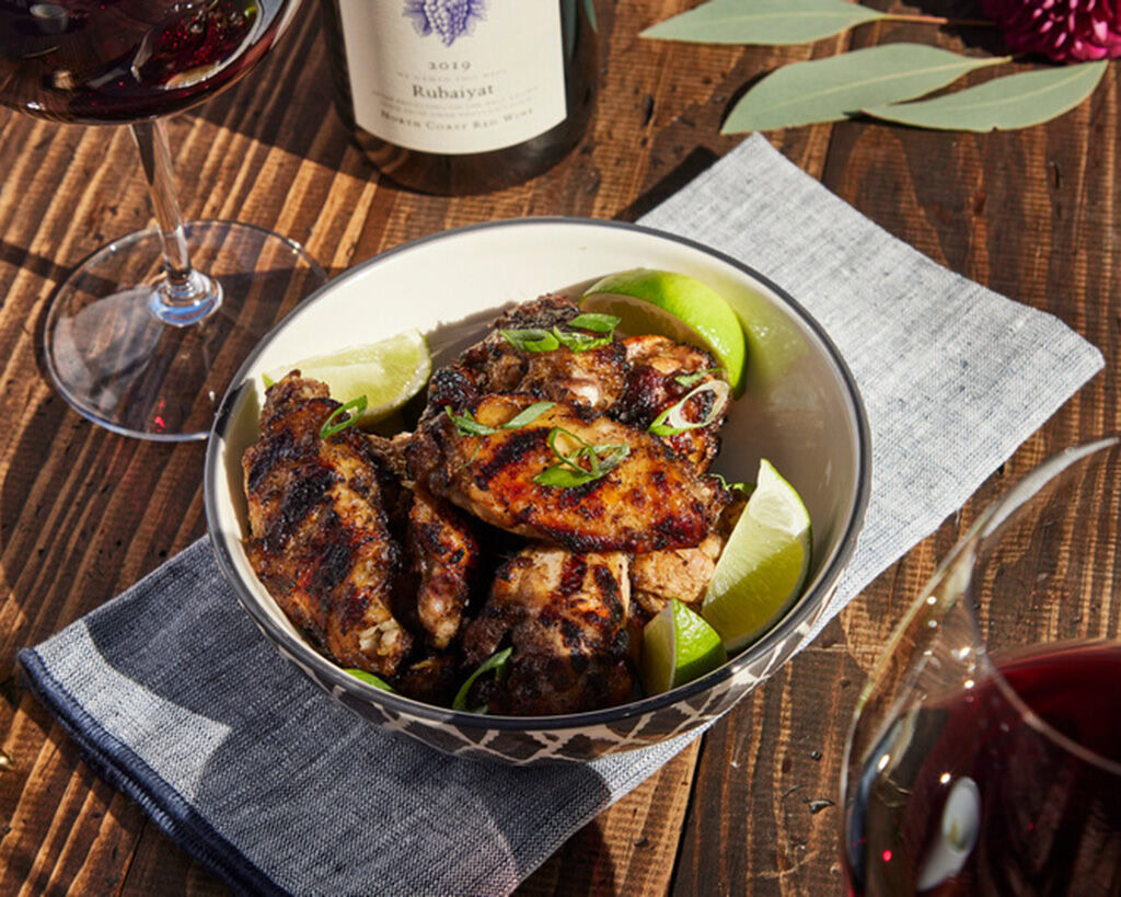 Grilled Jamaican Jerk Chicken Wings with lime wedges in a bowl on a wooden table, wine glasses, and a bottle of wine in the b.