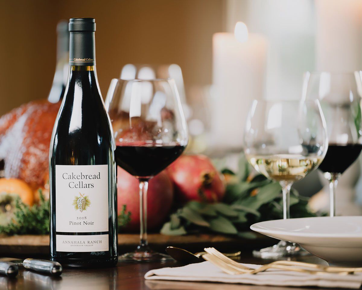 Bottle of Cakebread Cellars Pinot Noir with wine glasses and fresh fruit on a dining table.