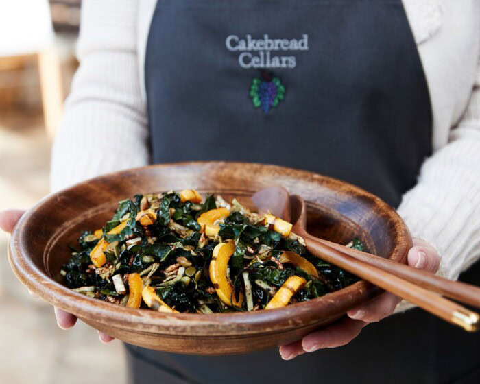 Kale and delicata squash salad served in a wooden bowl, held by a person wearing a Cakebread Cellars apron.