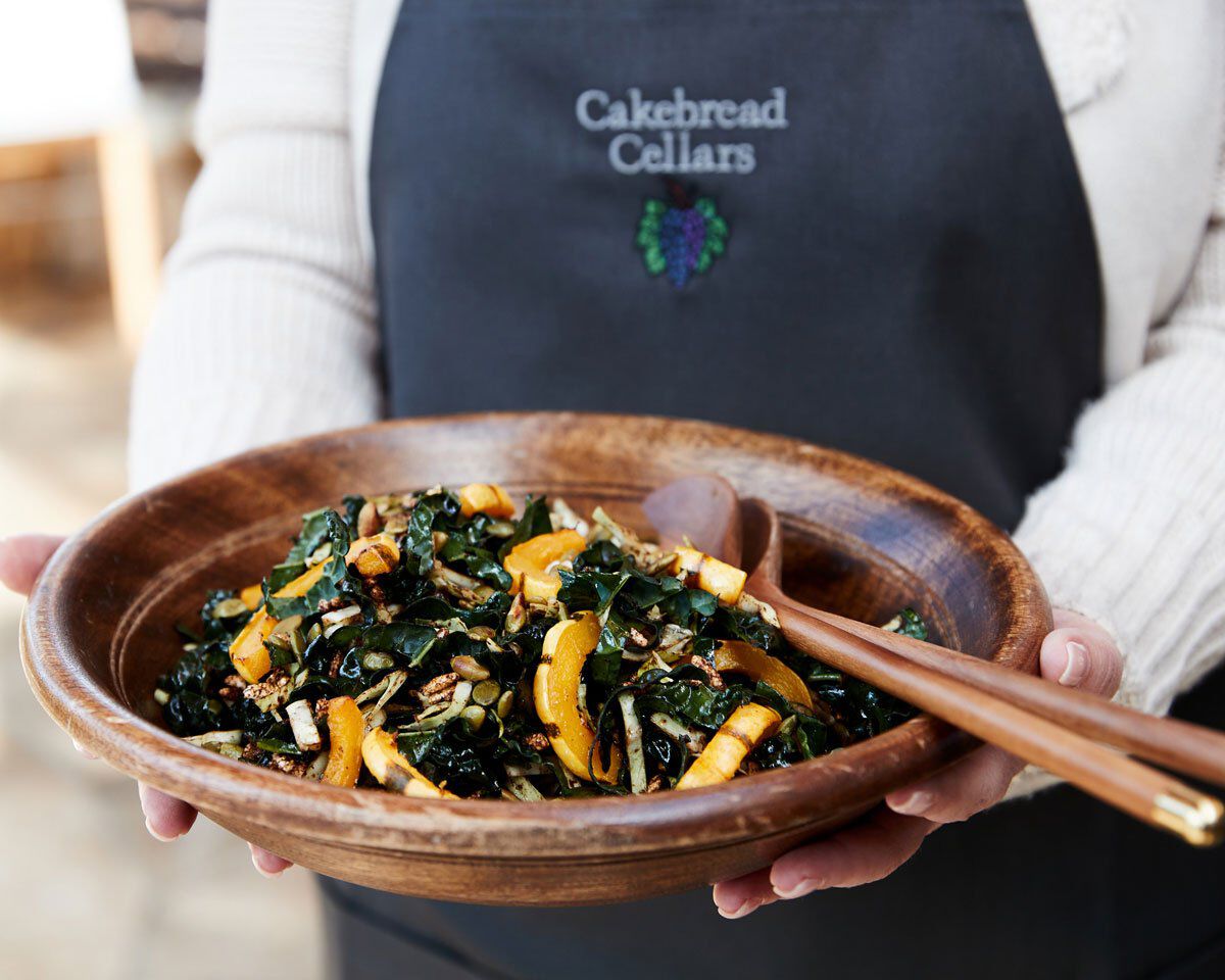 Kale and delicata squash salad served in a wooden bowl, held by a person wearing a Cakebread Cellars apron.