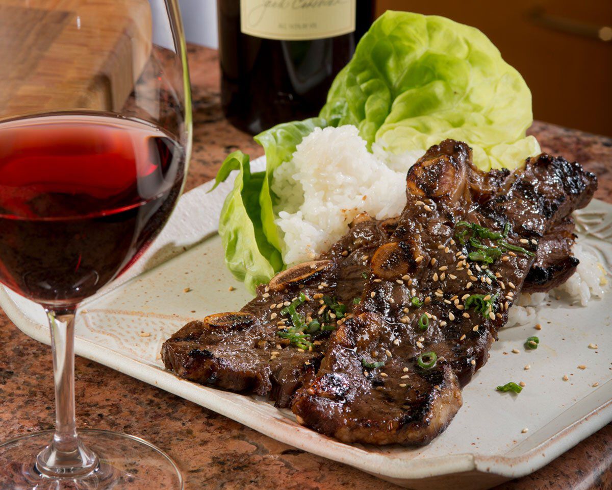 Grilled short ribs with lettuce, rice, and a glass of red wine on a white plate.