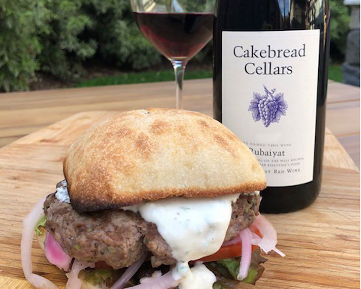 Lamb burger with whipped feta, pickled fennel, and fresh vegetables on a bun, with Cakebread Cellars wine in the background.