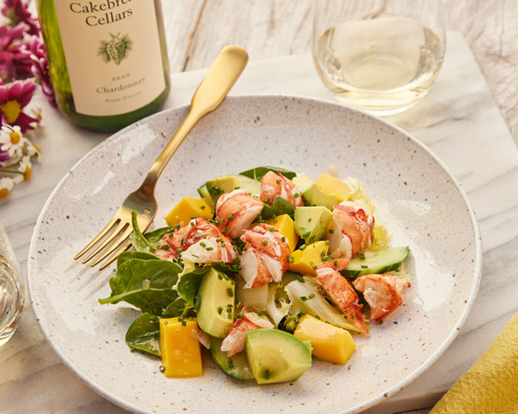 Lobster, avocado, and mango salad with honey-lime vinaigrette on a white plate, garnished with fresh greens, served with a bo.