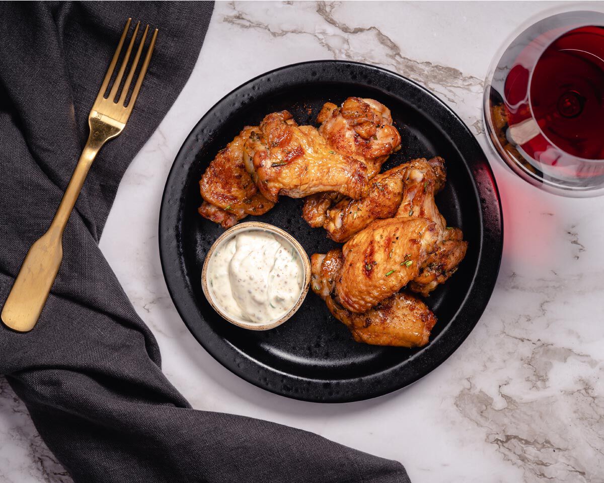 Juicy maple smoked chicken wings with herbed crème fraîche and a glass of Chardonnay salt on a black plate. Perfect pairing f.