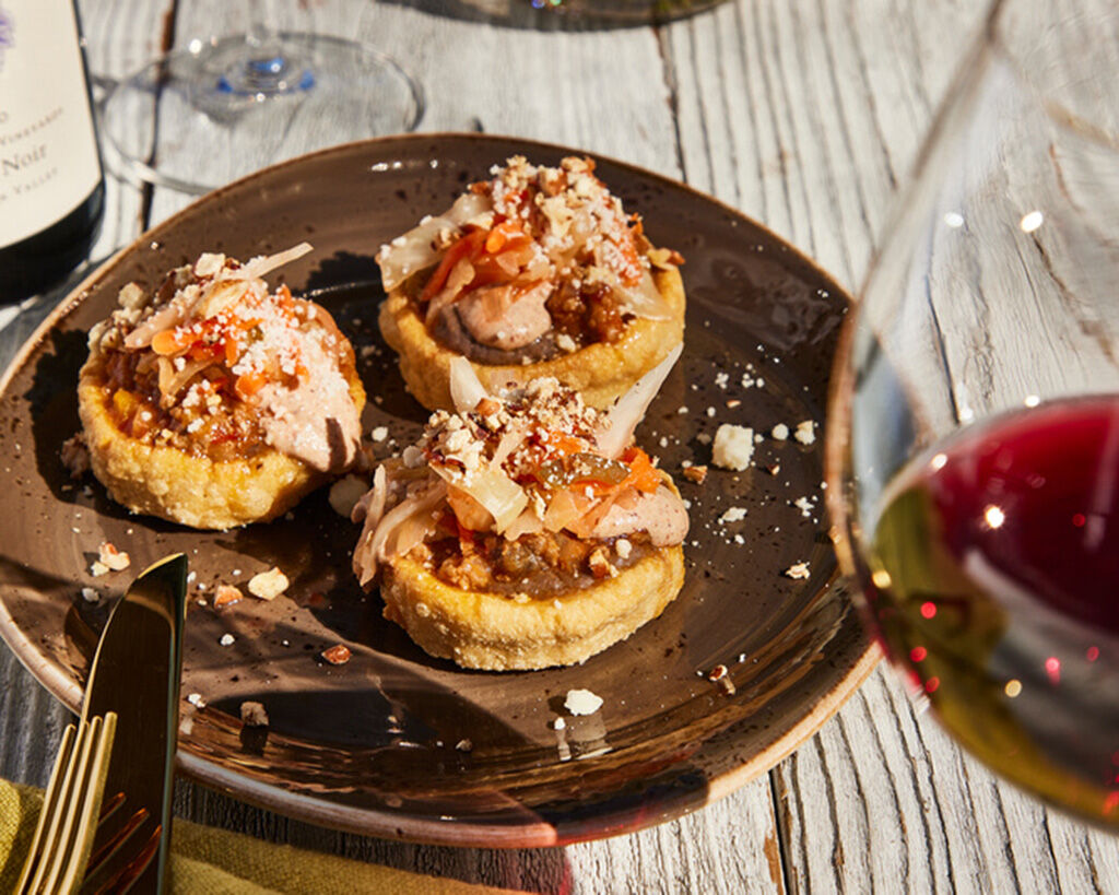 Mexican sopes topped with duck picadillo, pecan crema, and cheese on a dark plate, served with wine and rustic wooden table b.
