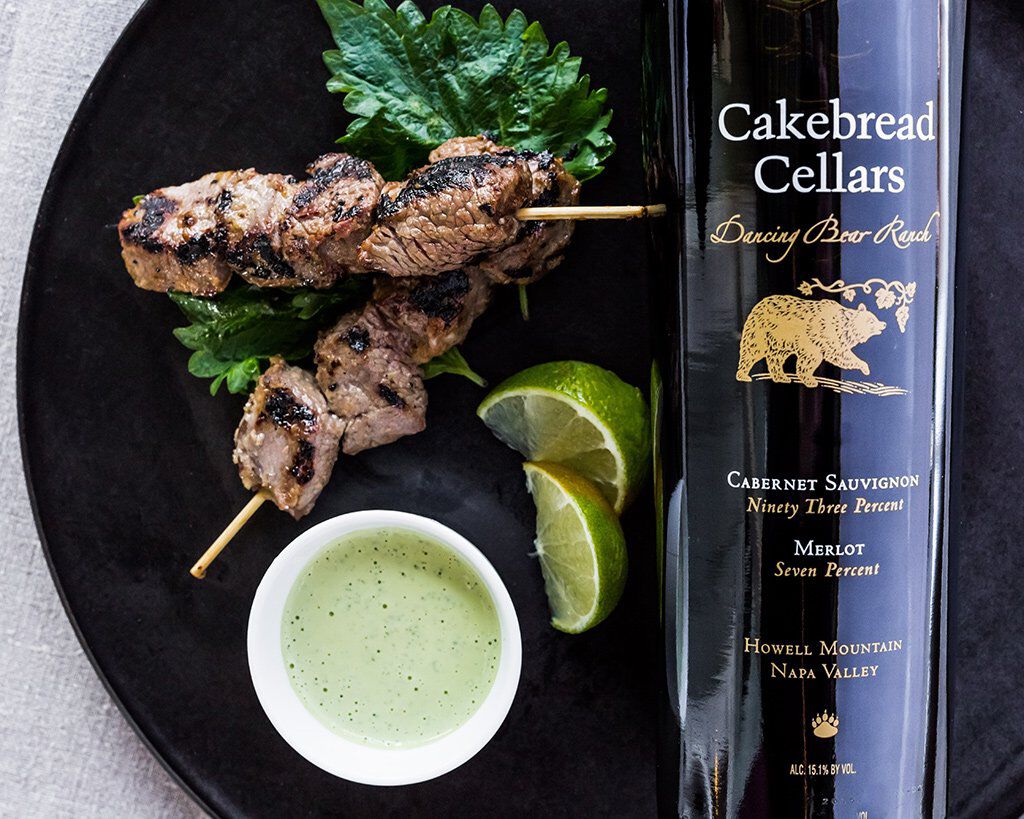 Succulent miso grilled beef brochettes with shiso aioli served with lime wedges and green dipping sauce on a black plate.