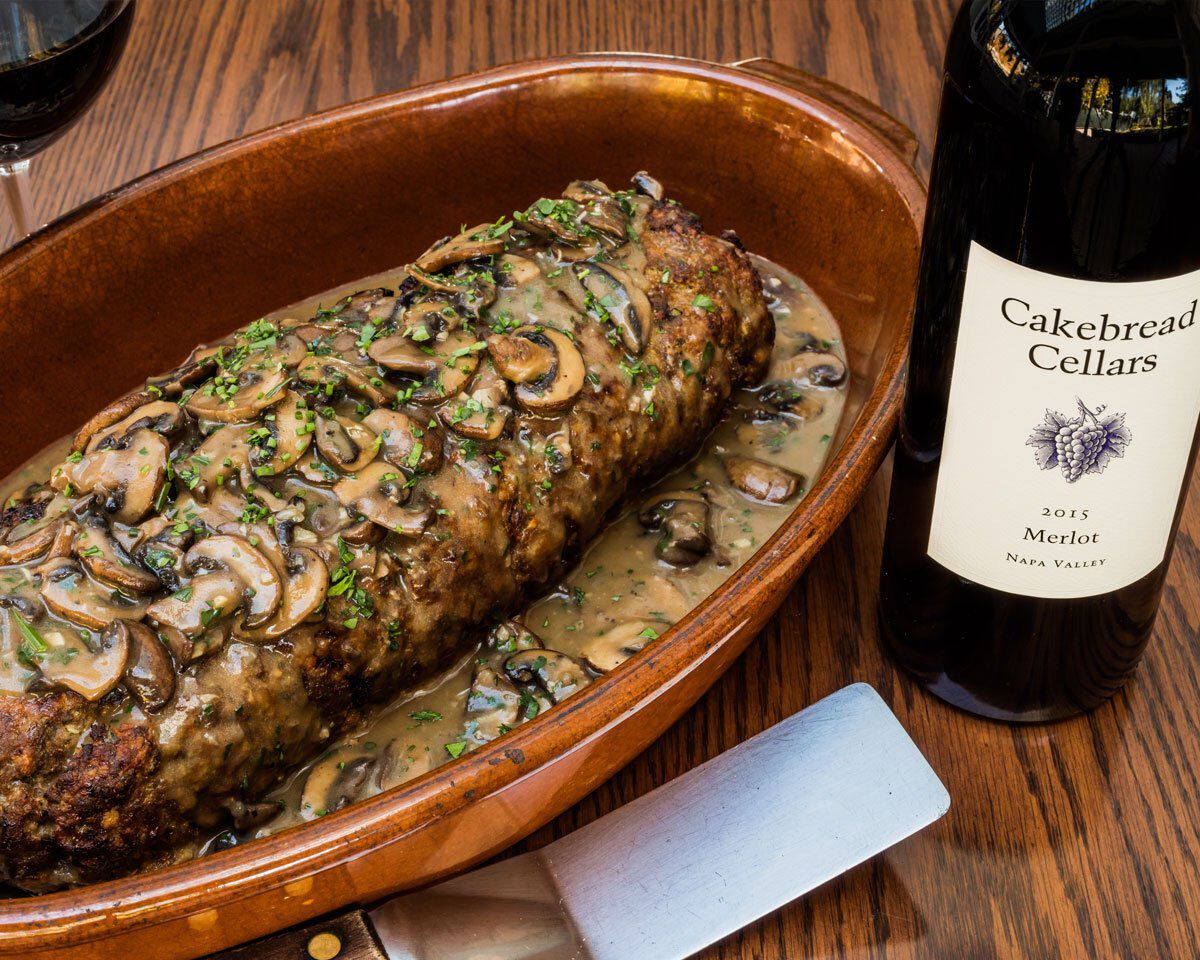 Mom's Meatloaf with Mushroom Gravy served in a wooden dish, with a bottle of Cakebread Cellars Merlot in the background.