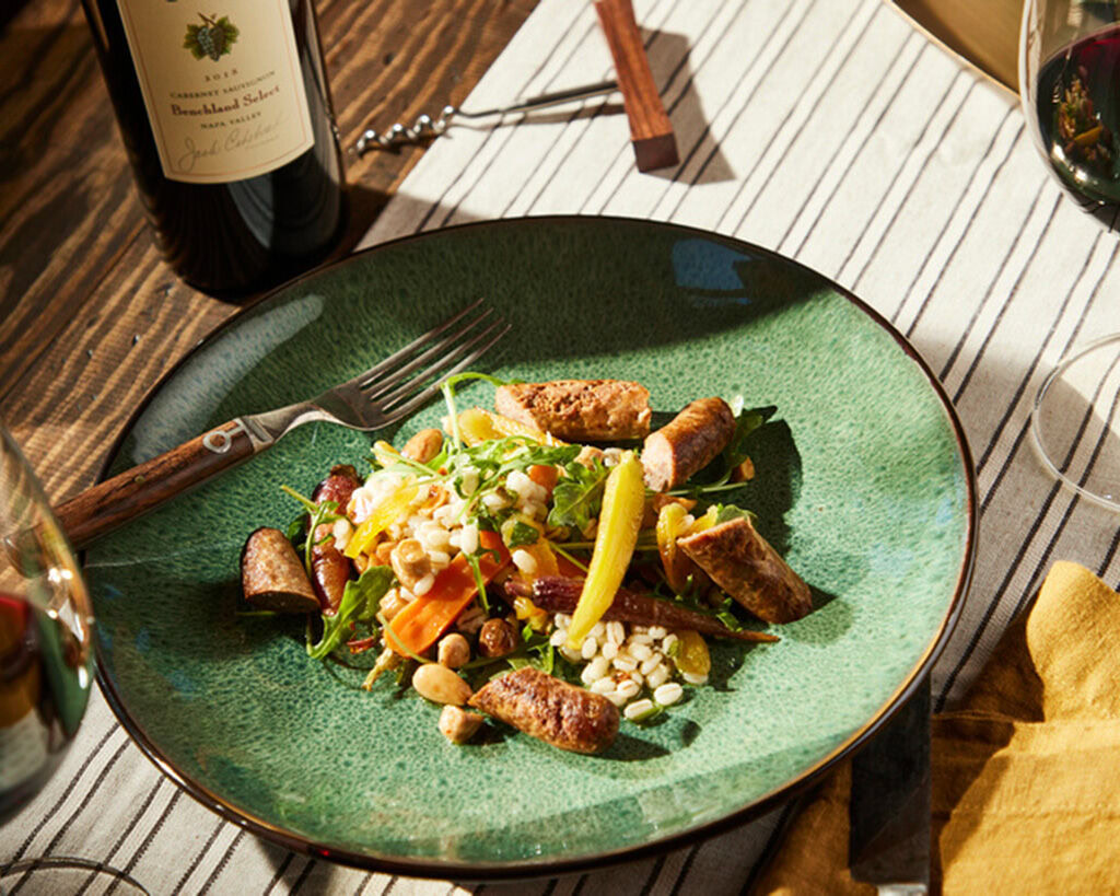 Grilled Moroccan lamb merguez sausage with roasted carrots, barley, dried apricots, and almonds on a green plate.