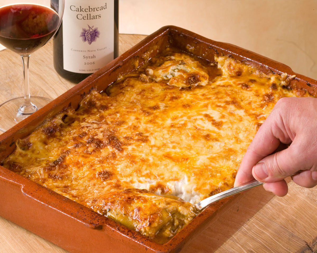 A delicious serving of Moussaka in a ceramic baking dish with a golden-brown cheese topping, accompanied by a glass of red wi.