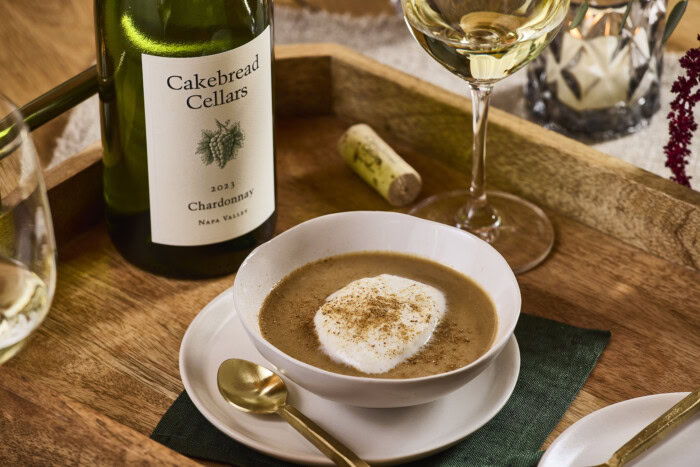 A lush bowl of mushroom cappuccino soup garnished with cream and spices, served on a wooden table with a bottle of Cakebread.