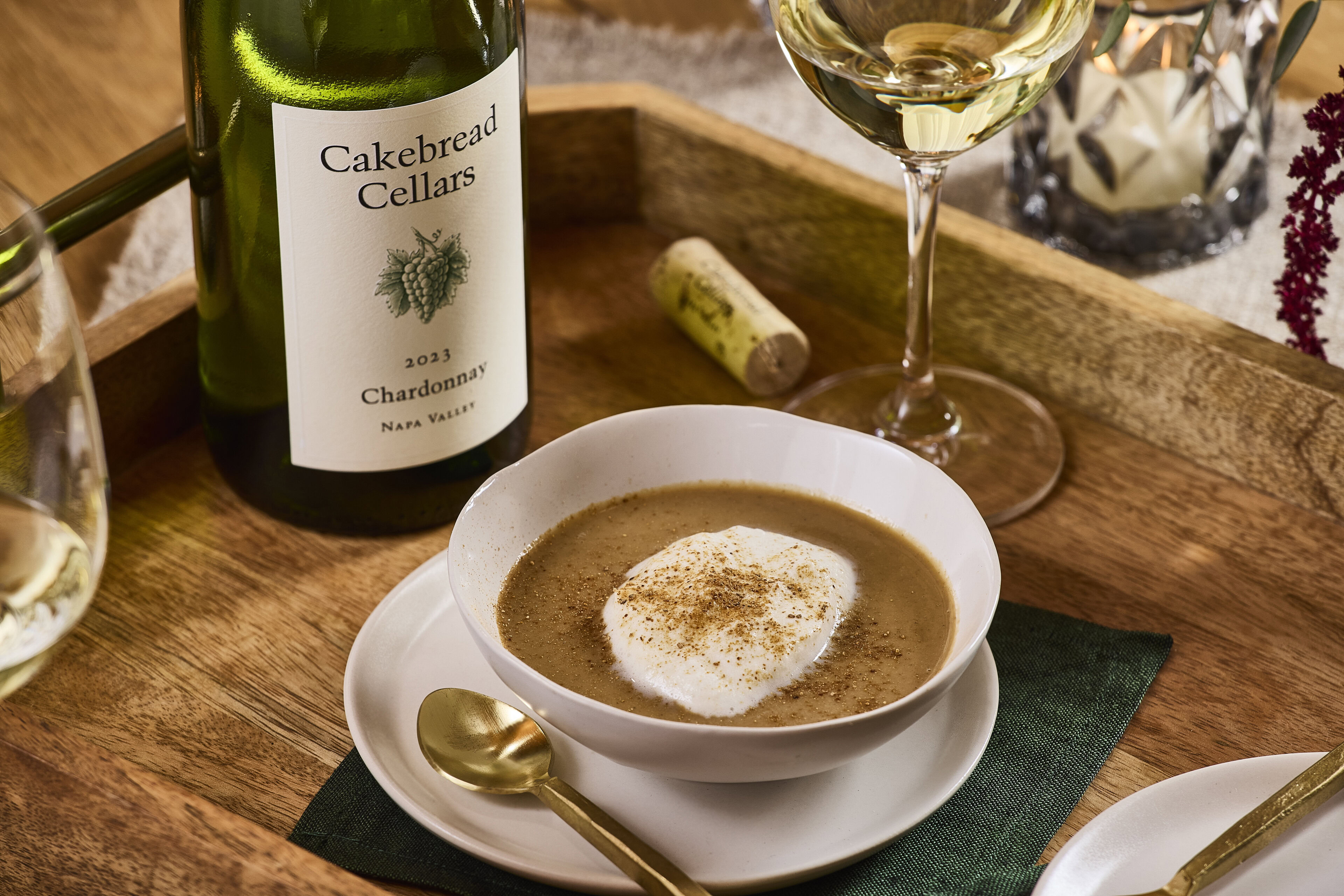 A lush bowl of mushroom cappuccino soup garnished with cream and spices, served on a wooden table with a bottle of Cakebread.