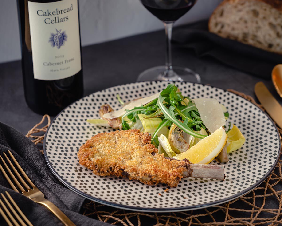 Grilled Parmesan crusted lamb chop with arugula and artichoke salad on patterned plate, wine glass, and bottle of Cakebread C.