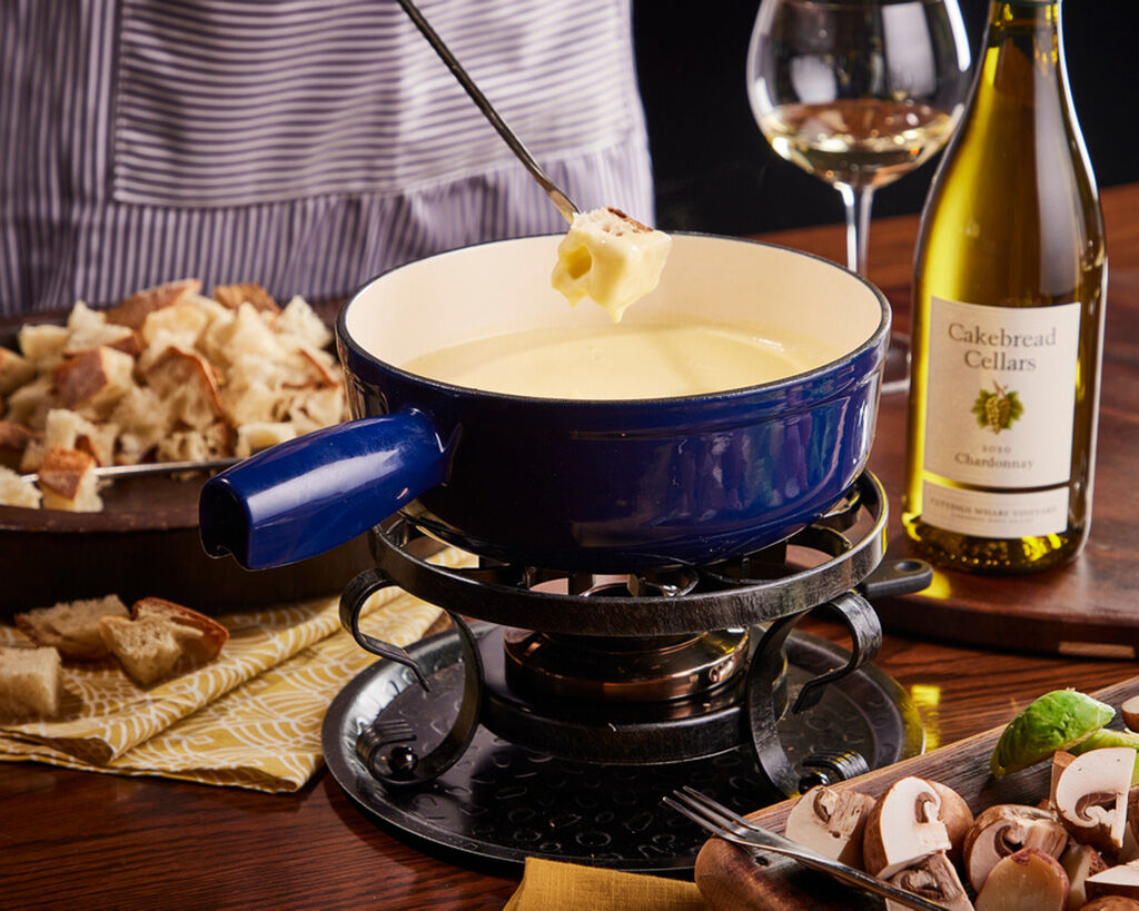 Creamy cheese fondue in a blue pot with a fondue fork, surrounded by bread, mushrooms, and a bottle of Cakebread Cellars wine.