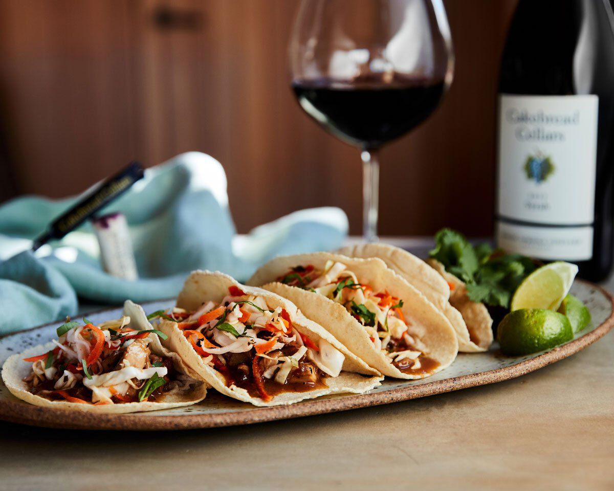 Pork adobo tacos with shredded meat, vegetables, and sauce on a ceramic plate, with wine and lime in the background.