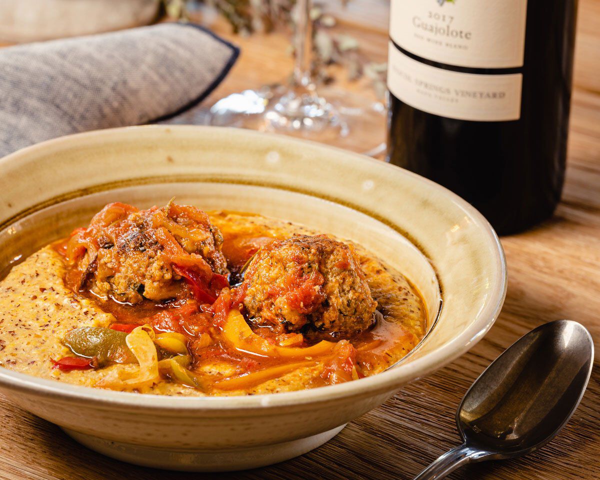 A colorful bowl of pork meatballs with braised peppers and onions in a creamy sauce, served on a rustic wooden table with a g.