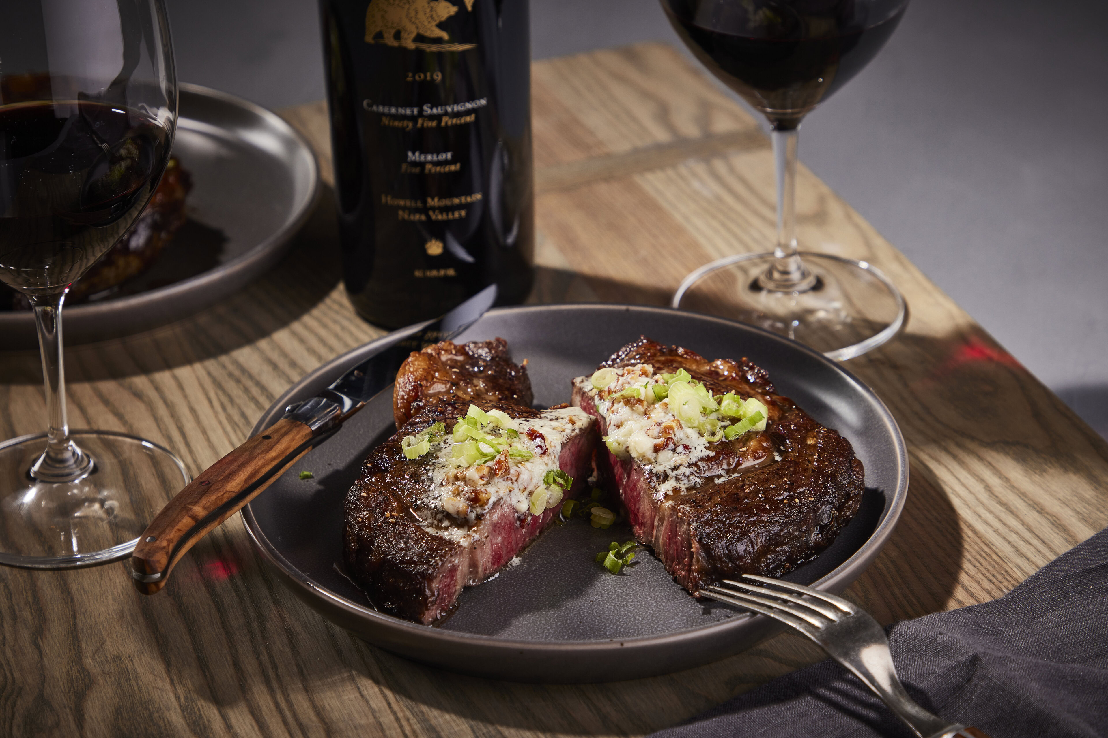 Juicy ribeye steak with pecan, bourbon, and blue cheese butter served on a black plate with wine glasses and a bottle in the.