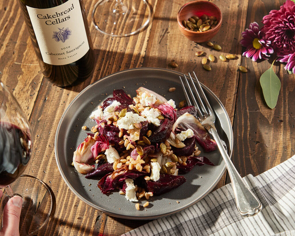 Roasted beet, radicchio, farro, and goat cheese salad on a dark plate with nuts and flowers, served with wine and fresh herbs.
