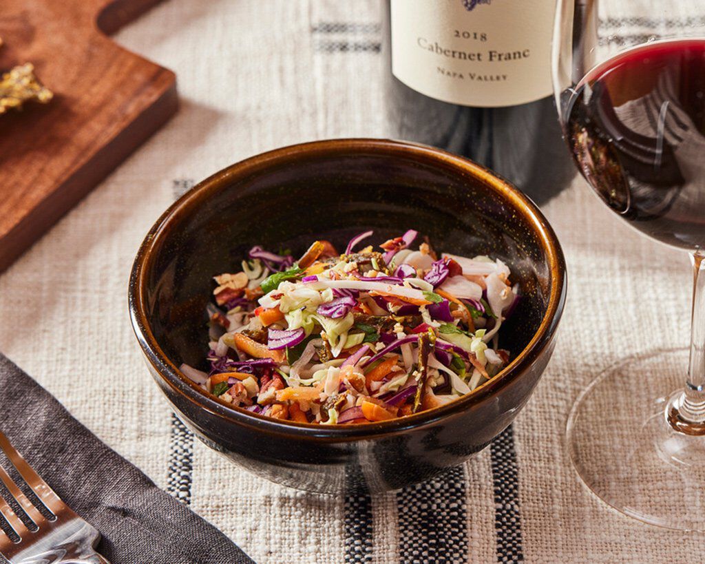 Bright Savoy cabbage slaw with fresh herbs, pecans, and vibrant vegetables in a dark bowl, paired with a glass of red wine an.