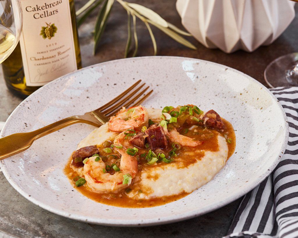 Shrimp and Andouille Sausage over creamy grits on a white plate, garnished with green onions, with a bottle of Cakebread Cell.